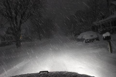 Truck headlights on an unplowed residential street before dawn during heavy lake-effect snow, snow streaking hard through the beams