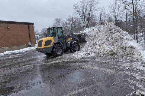 A compact wheel loader pushing a gray snow pile into the back corner of a commercial parking lot against a chain-link fence