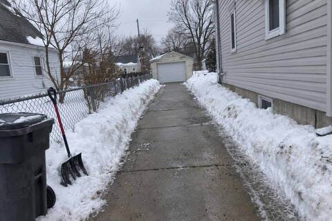 A long narrow driveway cleared to bare concrete running back between a chain-link fence and house siding to a detached garage at the rear of the lot, snow banked along both edges