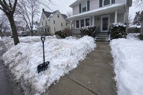 A public sidewalk in front of an older house cleared to bare concrete across its full width, snow walls on both sides and a shovel standing in the bank