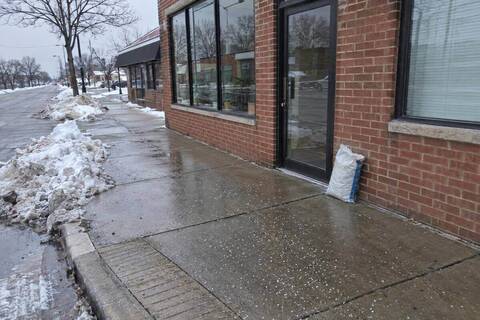 The sidewalk in front of a small brick storefront cleared to bare concrete across its full width, snow piled at the curb and the corner curb ramp cut clear