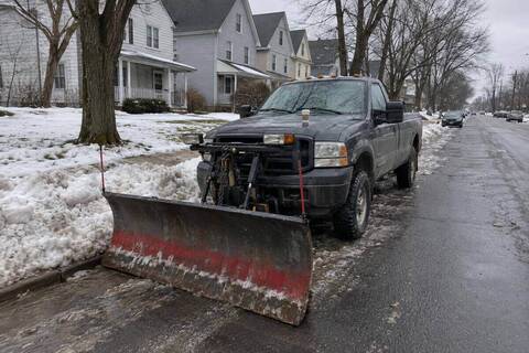 A pickup with a straight plow blade standing at the curb of a snow-covered residential street, blade lowered onto the pavement, plowed banks along the tree lawn