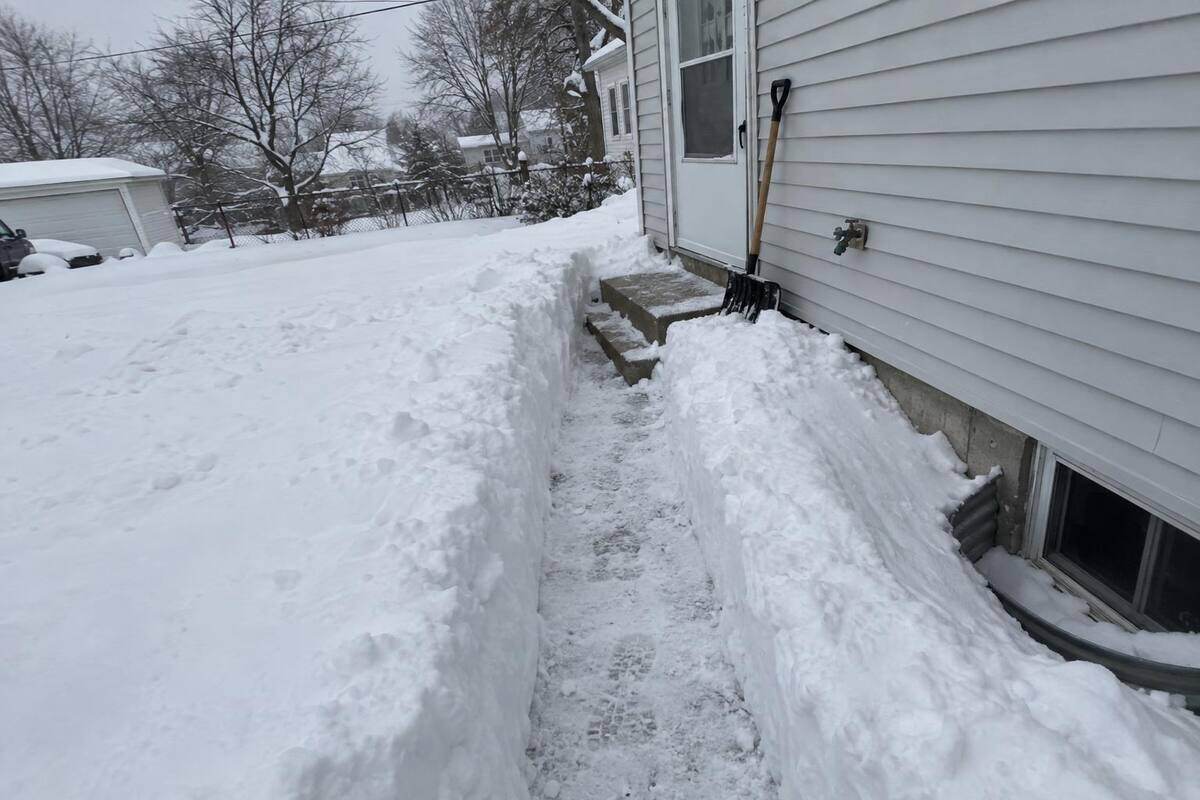 A shovel-width path cut across a deep snow-covered side yard to a side entry door with concrete steps, snow walls on both sides