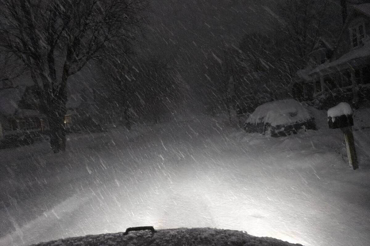 Truck headlights on an unplowed residential street before dawn during heavy lake-effect snow, snow streaking hard through the beams
