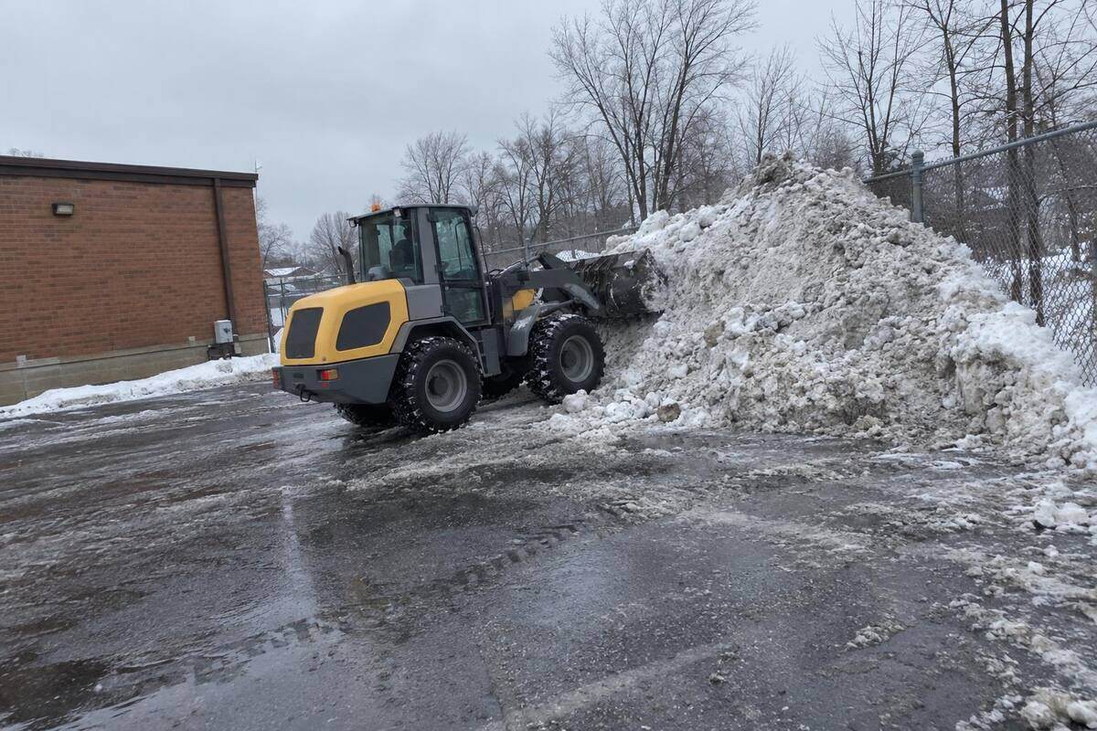 A compact wheel loader pushing a gray snow pile into the back corner of a commercial parking lot against a chain-link fence