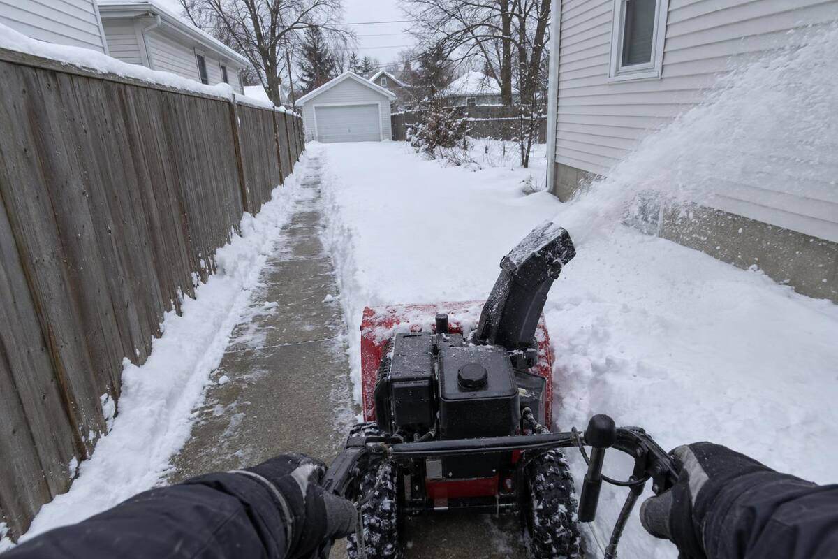 A walk-behind snowblower partway down a narrow driveway between a fence and house siding, chute turned to the side throwing snow onto the bank